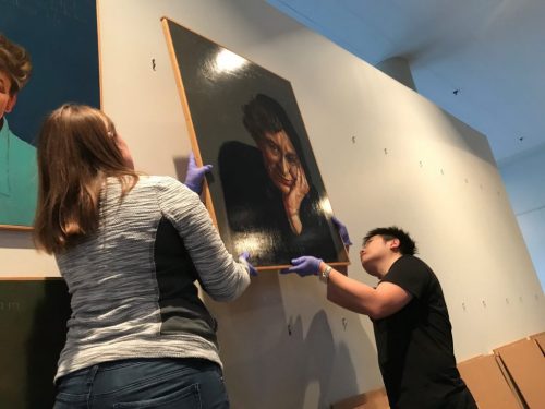 Museum studies students hang a painting on a wall.
